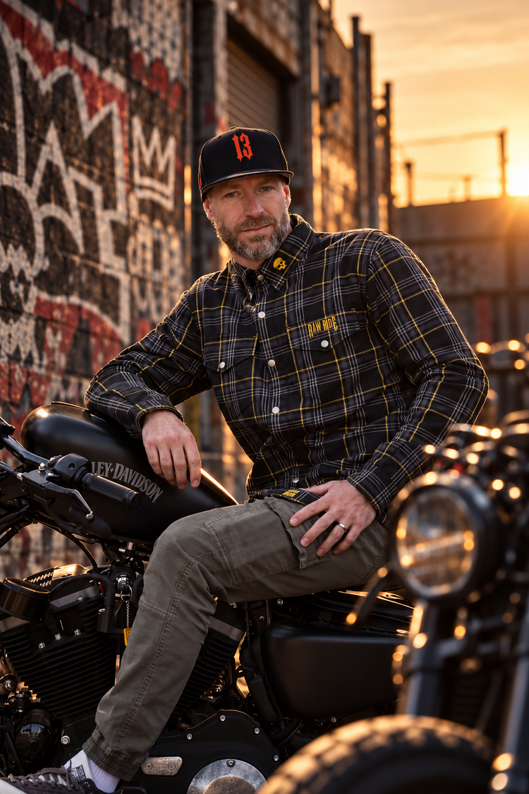 Homme assis sur une Harley-Davidson noire, en chemise kevlar RAW ARMOR noire à lignes jaunes et casquette 13, devant une forêt sombre.