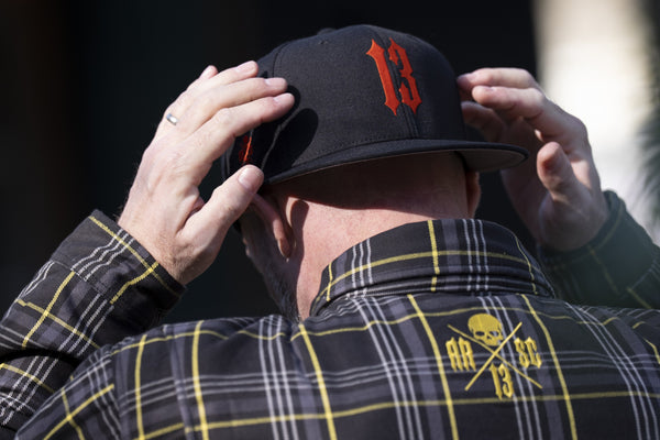 Homme de dos ajustant à deux mains sa casquette noire 13, en chemise kevlar à lignes jaunes au logo brodé dans le dos.