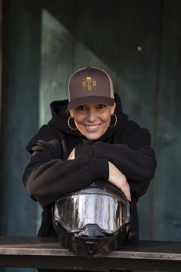 Femme souriante accoudée sur un casque de moto, en hoodie noir et casquette trucker brune au logo doré RR SC 13.