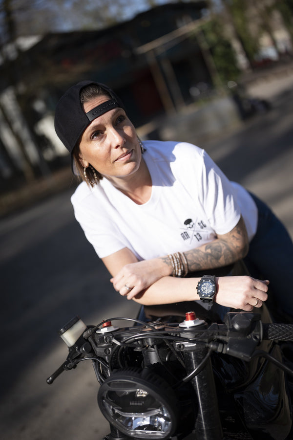 Femme accoudée au guidon de sa moto, en t-shirt blanc au petit logo Raw Ride et casquette noire portée à l'envers, le regard levé.