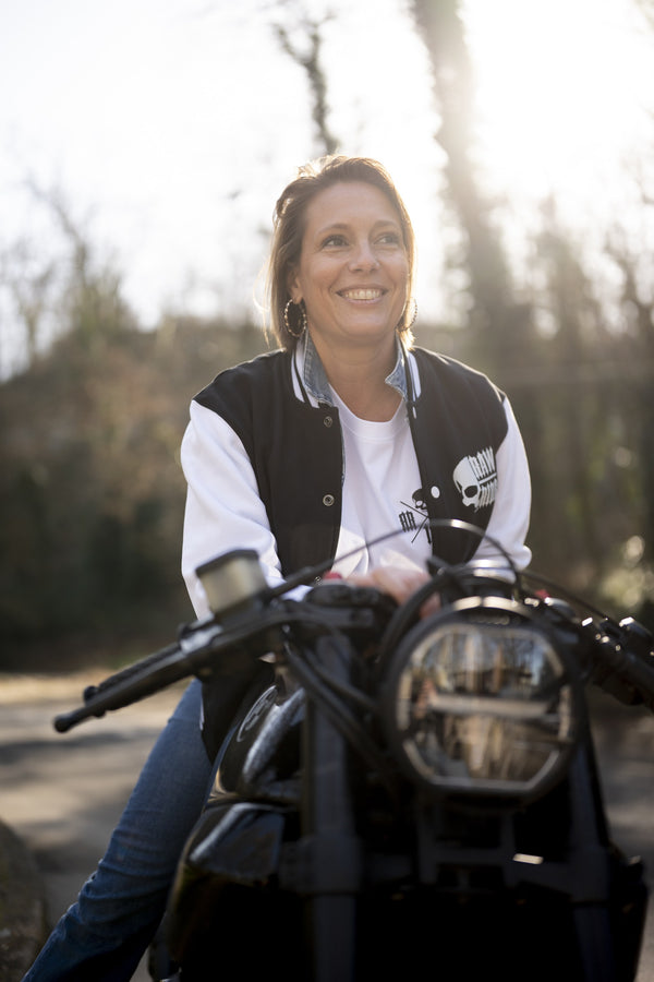 Femme souriante assise sur une moto en sous-bois, en veste collège noire à manches blanches Raw Ride, à contre-jour.