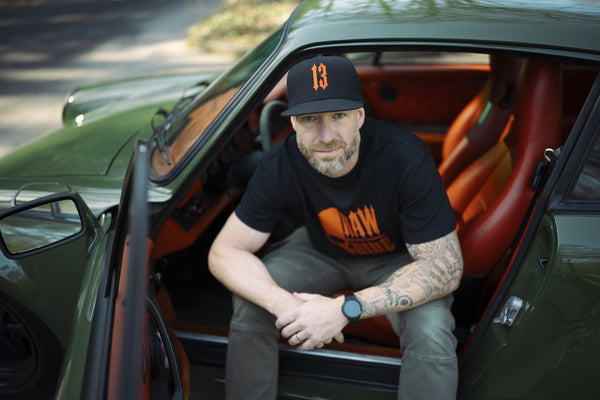 Homme assis sur le seuil d'une voiture ancienne verte aux sièges rouges, en t-shirt noir au crâne orange Raw Ride et casquette 13.