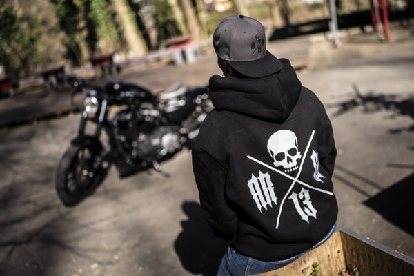 Personne de dos assise au soleil, en hoodie noir au grand logo crâne RR SC 13 et casquette grise, une moto noire en arrière-plan.
