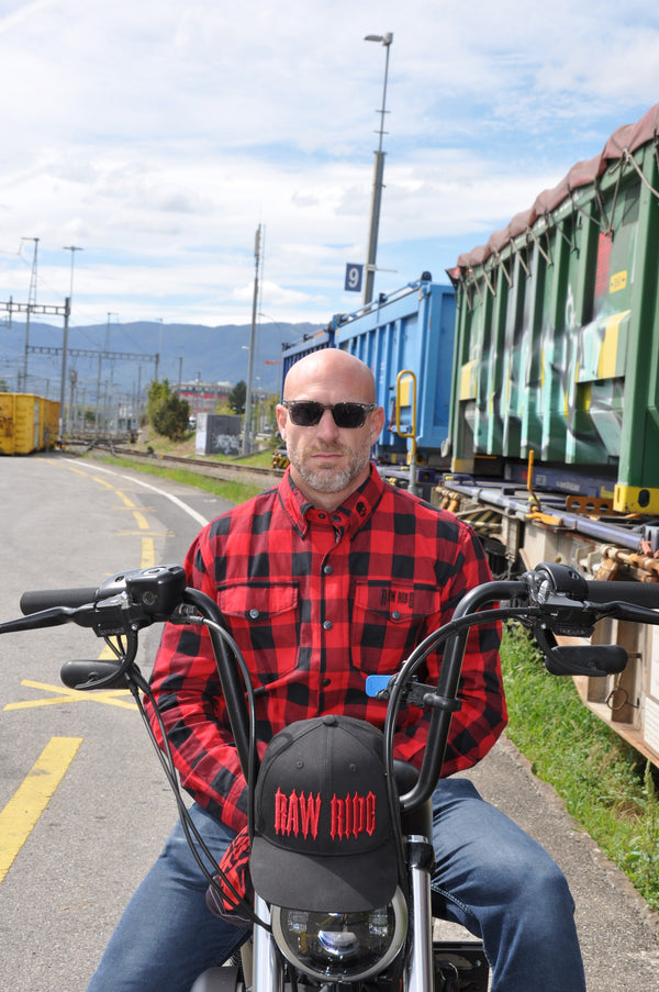 Homme de face assis sur sa moto entre deux trains de marchandises, en chemise flanelle rouge et noire, une casquette Raw Ride accrochée au phare.