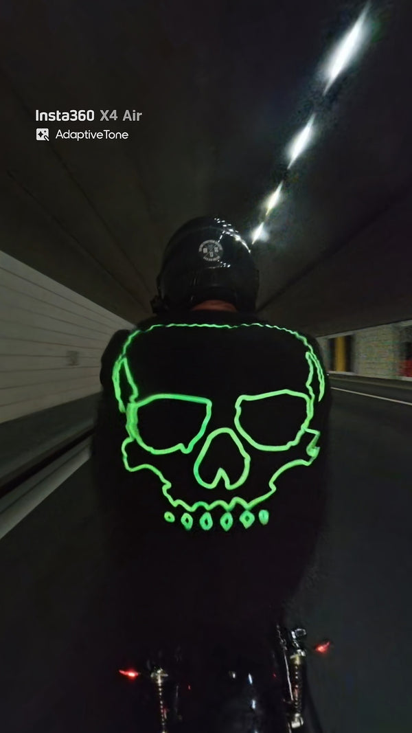 Motard vu de dos dans un tunnel, le grand crâne phosphorescent de son haut noir brille en vert.