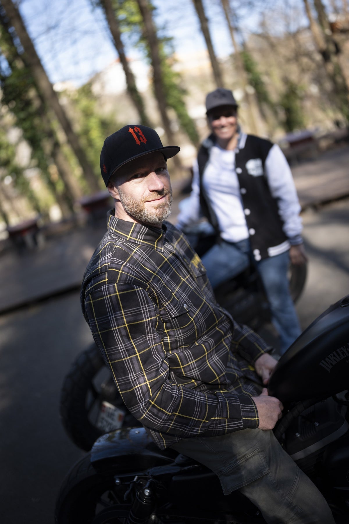 Homme en chemise kevlar à lignes jaunes assis sur sa moto, tourné vers l'objectif, et femme en veste collège Raw Ride debout derrière lui, dans un sous-bois.