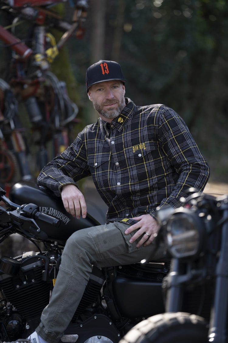 Homme assis sur une Harley-Davidson noire, en chemise kevlar RAW ARMOR noire à lignes jaunes et casquette 13, devant une forêt sombre.