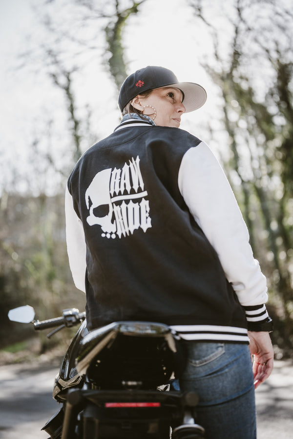 Femme de dos sur une moto, tête tournée vers la droite, en veste collège noire à manches blanches avec le grand logo crâne Raw Ride dans le dos.
